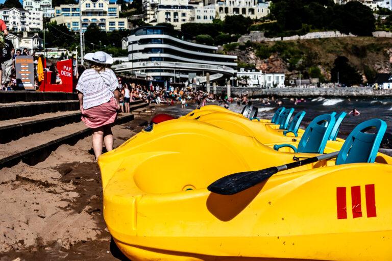 Row of yellow pedal boats on the sand with people on Torre Abbey beach behind