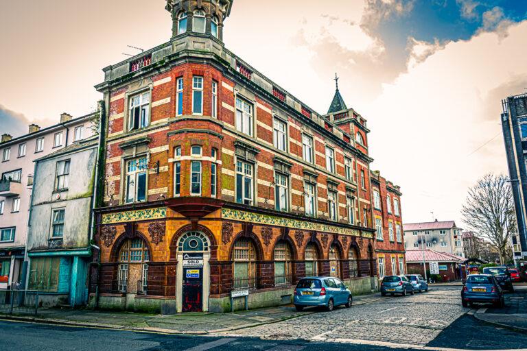 Ornate red-brick corner building on a cobbled street with parked cars and a cloudy sky.