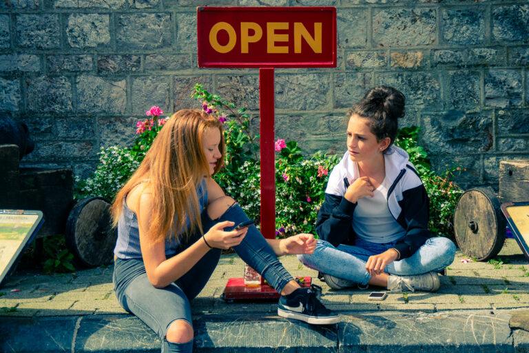 Two people sitting on the pavement talking beneath an “OPEN” sign by a stone wall.