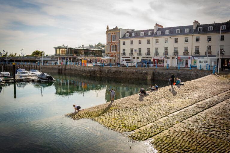 Torquay Harbour waterfront with boats moored in calm water and people on a cobbled slipway.