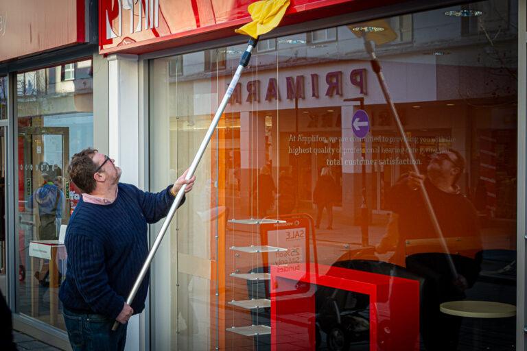 Person using a long pole to clean a glass shopfront window, with reflections of the street in the glass.